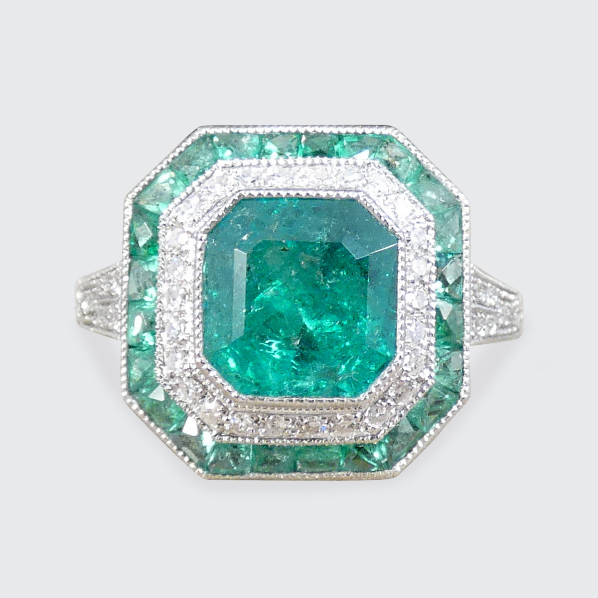 Striking Art Deco-style ring featuring a central emerald-cut emerald, surrounded by a double halo of calibre-cut emeralds and pavé-set diamonds, all set in platinum.