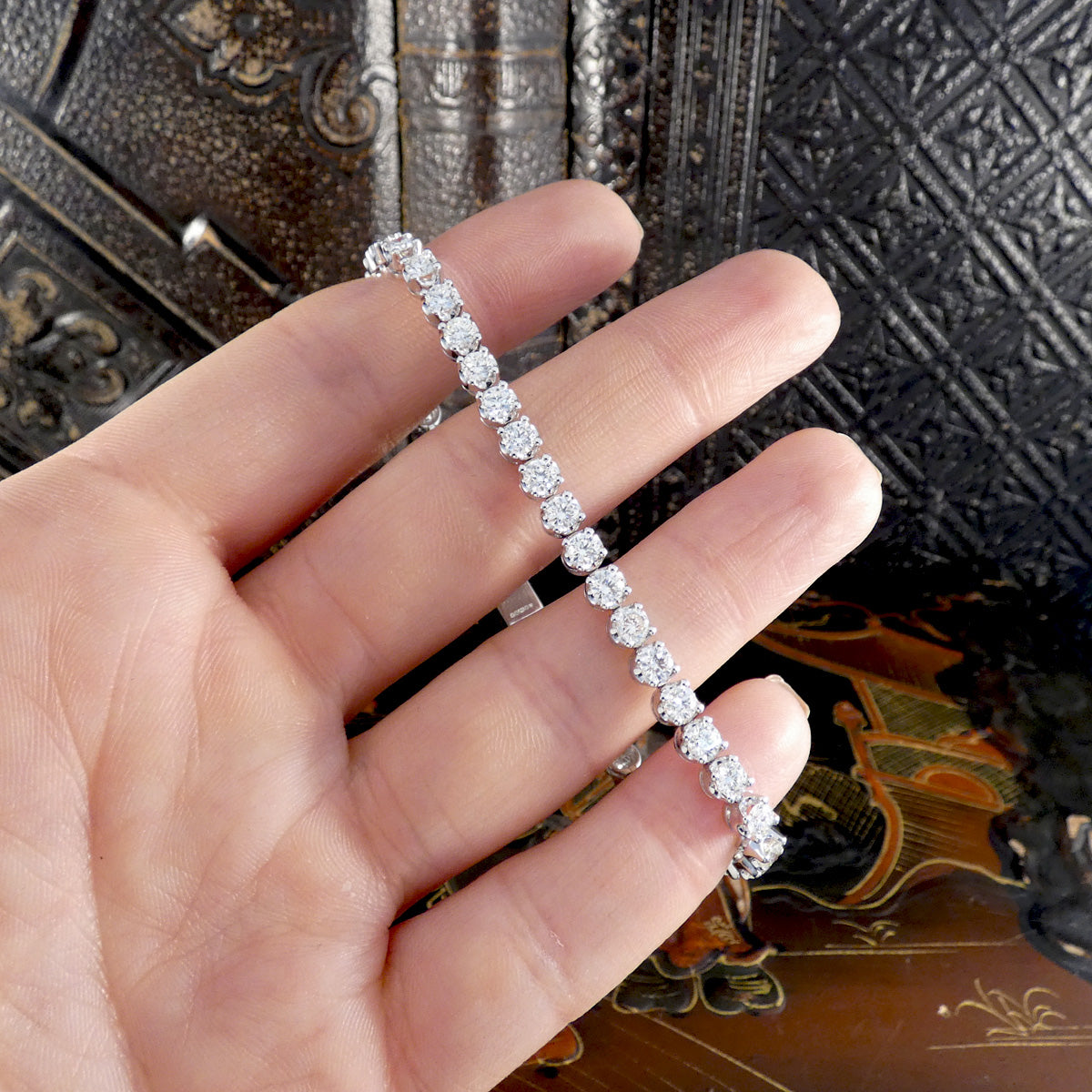 Hand holding a white gold tennis bracelet set with round brilliant-cut diamonds in four-claw settings, displayed against an ornate dark background.