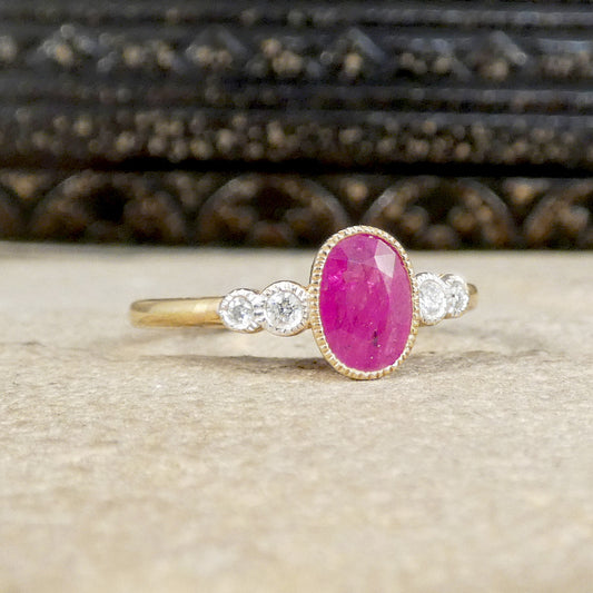 A vintage-style yellow gold ring featuring an oval pink ruby gemstone in a beaded halo, flanked by two small round clear stones on each side, resting on a textured beige surface with a dark carved wooden background.