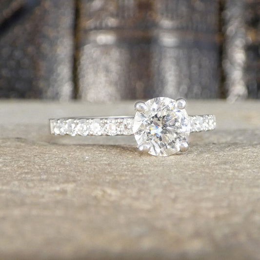 A solitaire diamond engagement ring with a mirco claw set band, resting on a textured stone surface with a blurred dark background.