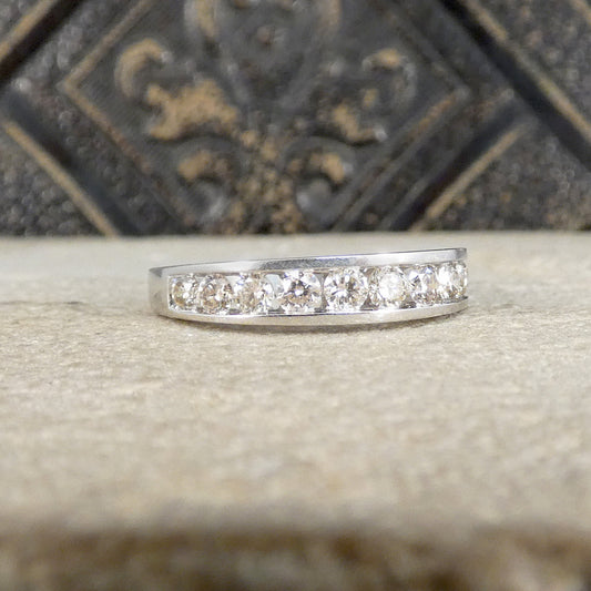A silver band ring featuring a row of clear round-cut gemstones, displayed on a textured beige surface with a dark, ornate background.