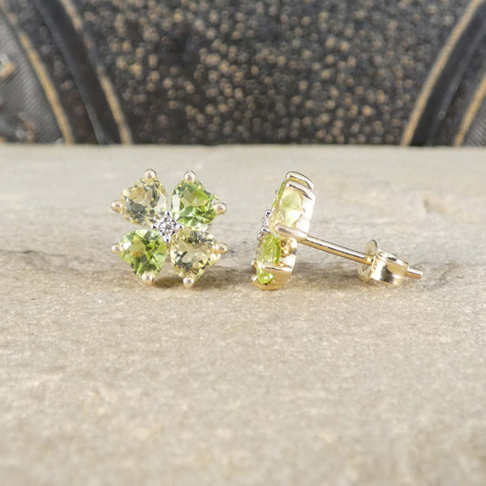 A pair of yellow gold stud earrings with four green gemstones arranged in a flower shape, resting on a textured beige stone surface with a dark patterned background.