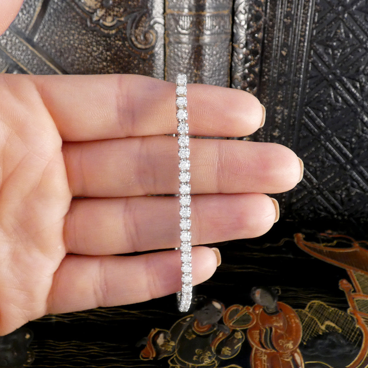 Hand holding a sparkling diamond tennis bracelet, showcasing a continuous line of round brilliant-cut diamonds set in white gold, against a decorative antique background.