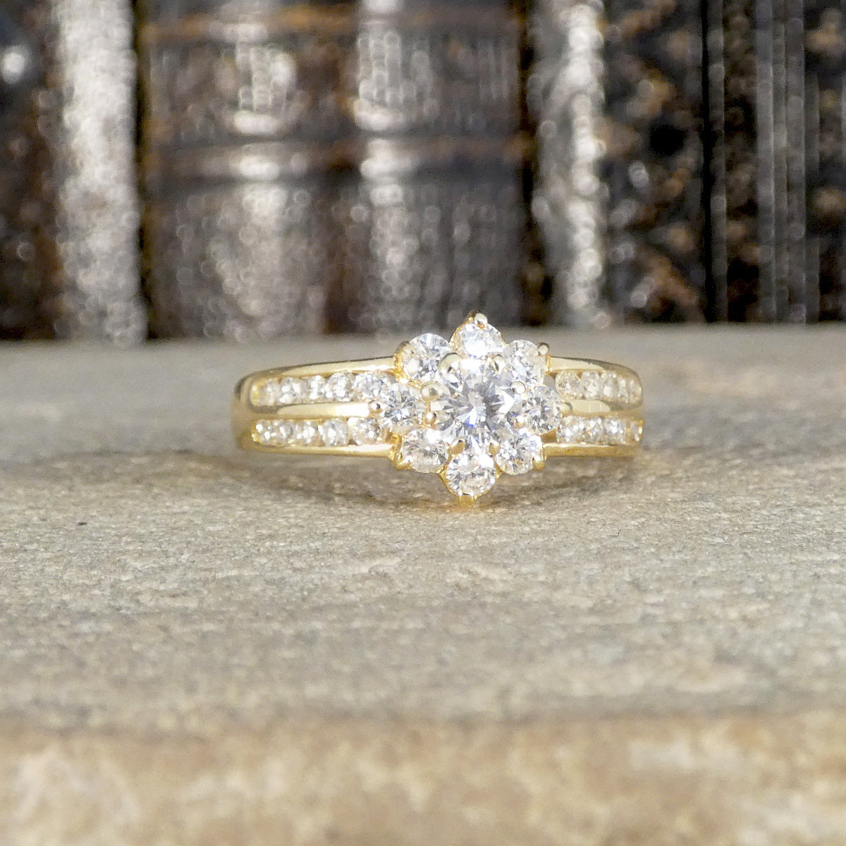 Dazzling diamond cluster ring in 18ct yellow gold with a floral-inspired design and diamond-set split shoulders, displayed on a textured stone surface.