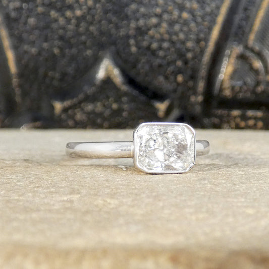 Radiant cut bezel set solitaire diamond ring in 18ct white gold, displayed on a textured surface with an ornate black and gold background.