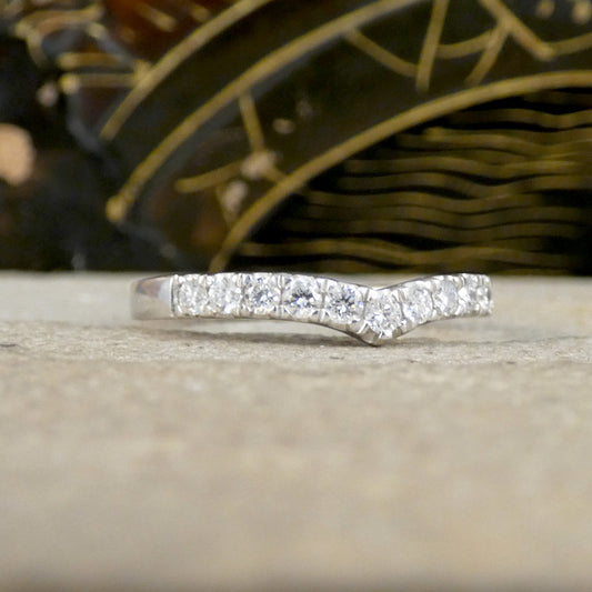 A silver band ring featuring a wavy, chevron-like arrangement of clear pavé-set gemstones, resting on a light beige textured surface with a dark, patterned background.