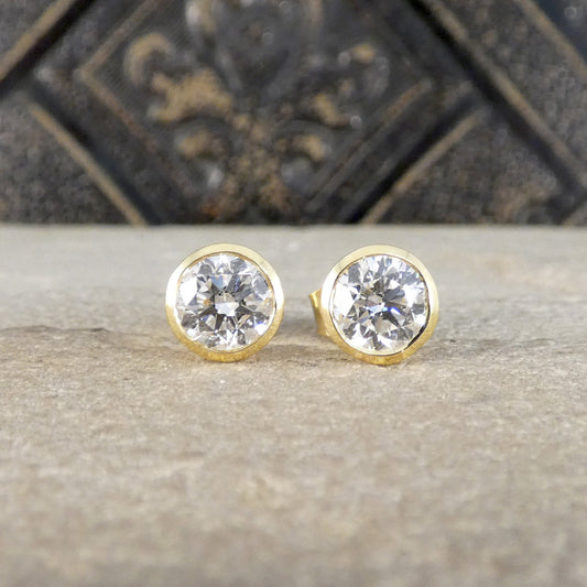 A pair of round gold stud earrings with clear faceted gemstones, displayed on a textured stone surface with a dark decorative background.