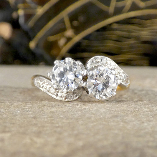 A silver-tone ring featuring two large round-cut clear gemstones and smaller accent stones along the curved band, resting on a textured stone surface with a blurred dark background.