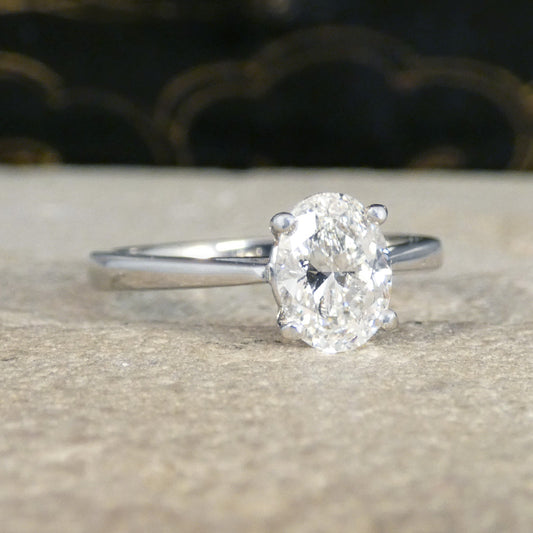 An oval-cut diamond solitaire ring in a silver-toned setting, resting on a textured beige stone surface with a dark, blurred background.