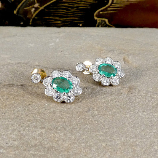 A pair of silver-toned cluster earrings with oval green emerald-cut center stones surrounded by small clear stones, displayed on a textured stone surface.