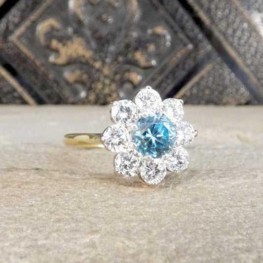 A flower cluster ring featuring a central round-cut light blue gemstone surrounded by clear round stones, set on a yellow gold band, displayed on a textured stone surface.