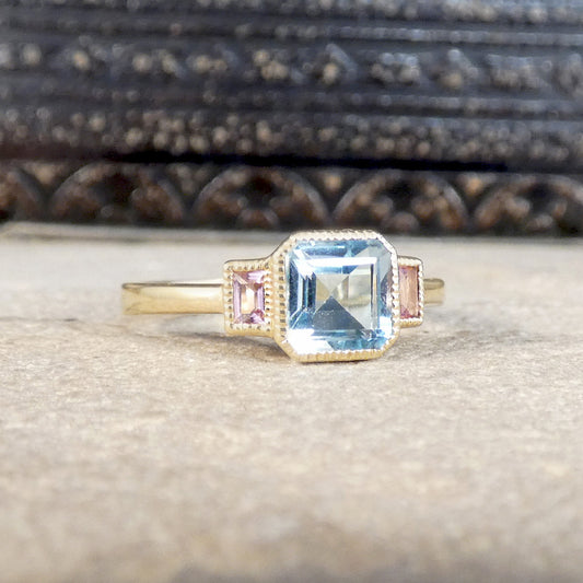 A gold ring featuring a large emerald-cut light blue center stone and two small pink side stones, displayed on a textured beige surface with a dark carved wooden background.