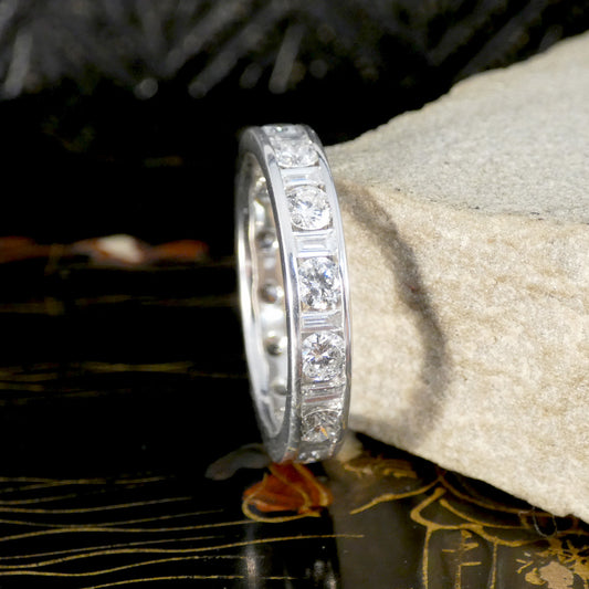 Close-up of a white gold eternity ring featuring alternating round brilliant and baguette-cut diamonds in a channel setting, elegantly displayed against a natural stone and decorative background.