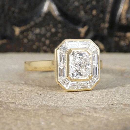A yellow gold ring featuring an octagonal bezel-set diamond with baguette side stones, resting on a textured stone surface with a dark, blurred background.