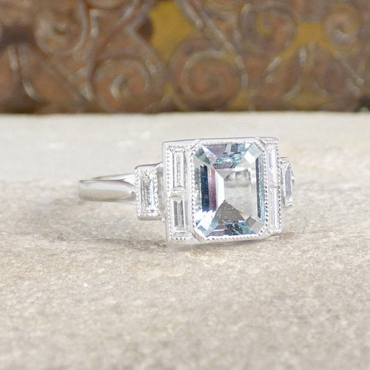 A gorgeous Emerald cut Aquamarine in a rub over bezel setting with a millrgain detailed edge. Accompanied by Diamonds in an Art Deco style this lovely ring is set in Platinum.