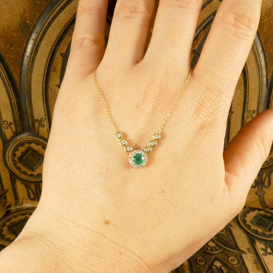 A stunning new necklace crafted in 18ct yellow gold, featuring a vibrant green emerald centre piece surrounded by a halo of sparkling diamonds, complemented by additional bezel-set diamonds in an illusion setting along the chain. Displayed on hand for size ratio.