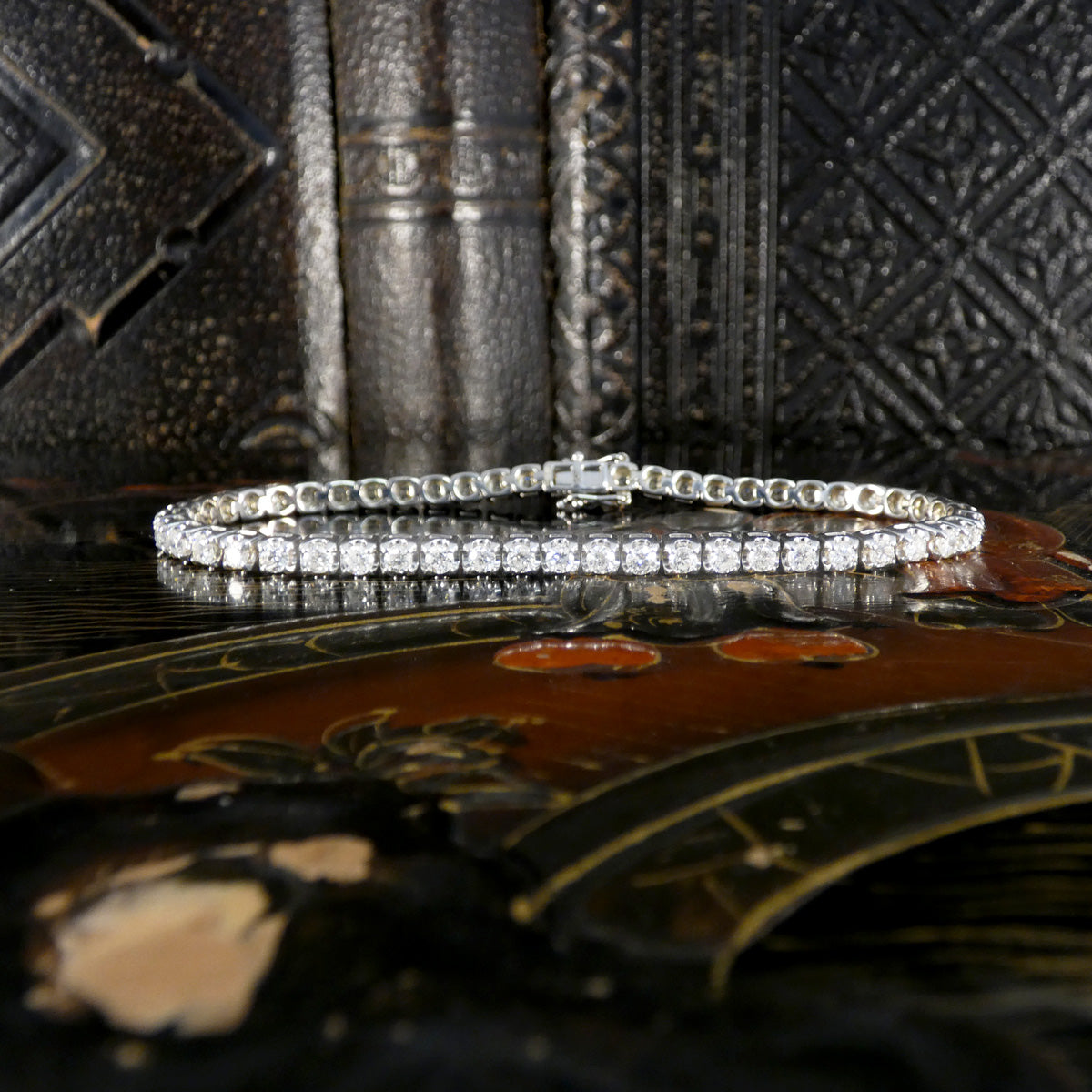 Elegant diamond tennis bracelet laid flat to display its uniform row of round brilliant-cut diamonds set in white gold, placed on an ornate lacquered surface with an antique book backdrop.