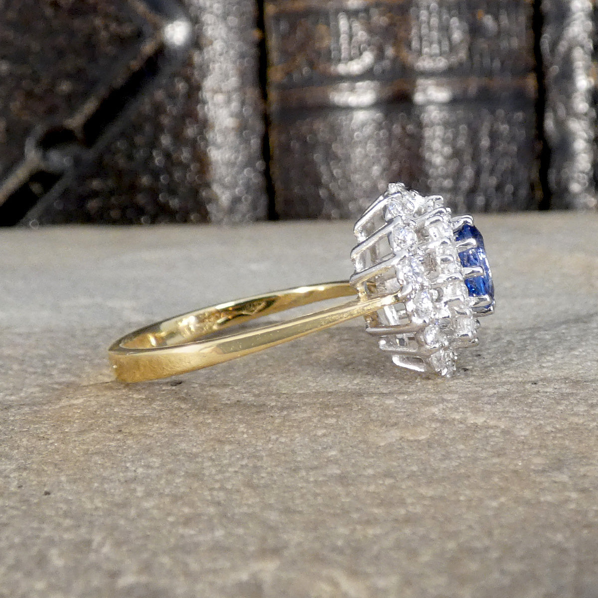 Side profile of Vintage-style cluster ring featuring a central oval blue sapphire surrounded by multiple round brilliant-cut diamonds, set in white and yellow gold.