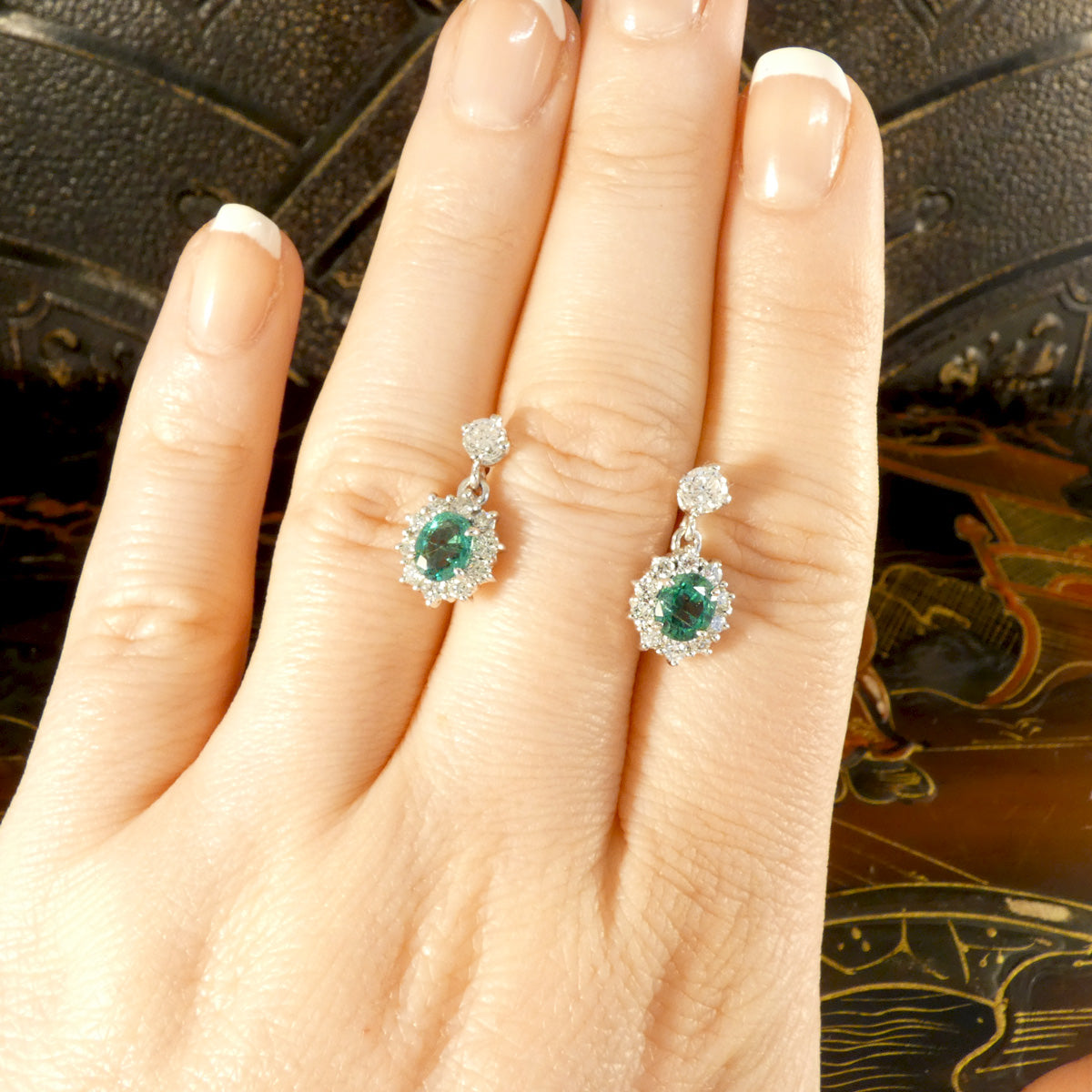 A beautiful pair of Oval Cut Emeralds surrounded by a halo of bright and sparkly diamonds hanging from a diamond stud as a drop earrings. Fully crafted in 18ct White gold, making the perfect gift. Displayed on hand for size and colour reference.