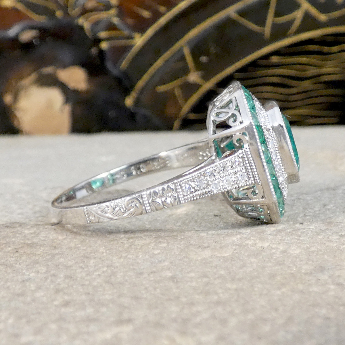Side view of Striking Art Deco-style ring featuring a central emerald-cut emerald, surrounded by a double halo of calibre-cut emeralds and pavé-set diamonds, all set in platinum. Showing detail down the shoulder of the ring in 850 Platinum.
