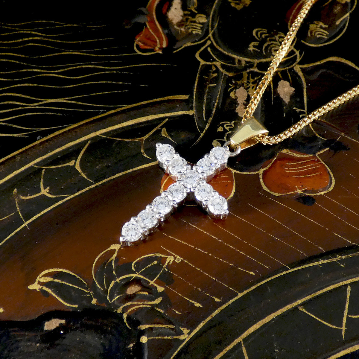 Elegant diamond cross pendant lying on a lacquered decorative surface, featuring round brilliant-cut diamonds set in 18ct white gold with a polished yellow gold bail and matching chain.