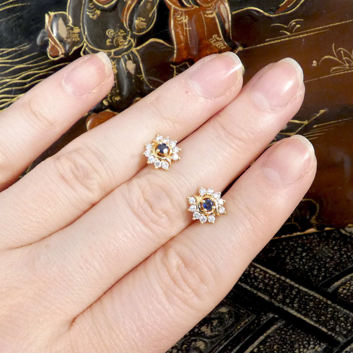 Gold earrings with blue Sapphire gemstones and a diamond cluster on a hand against an ornate background