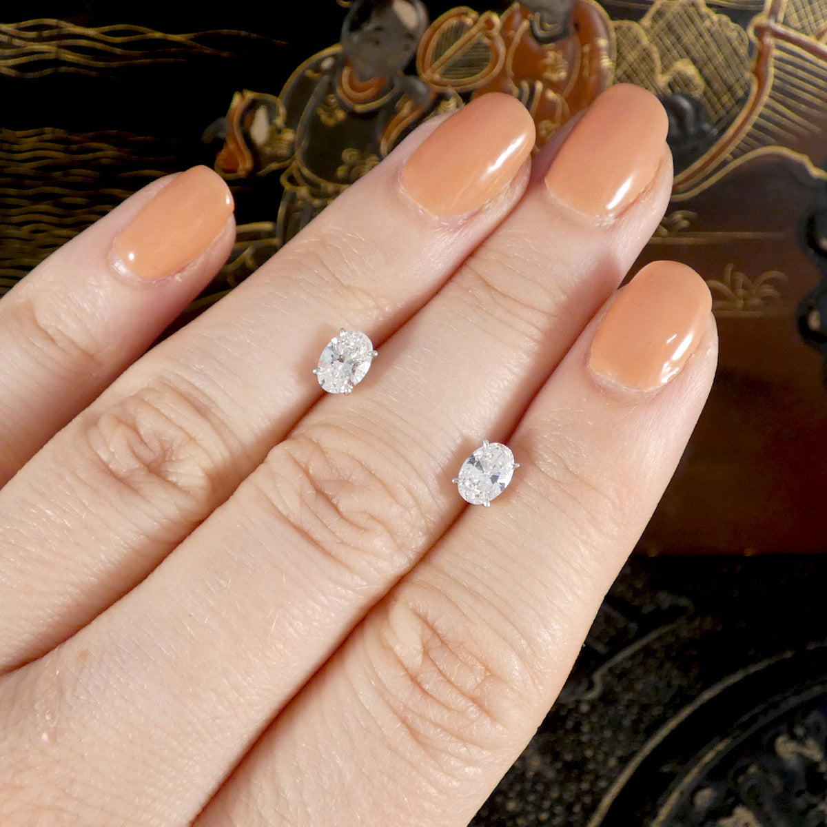 Diamond earrings on a hand with peach nail polish against a decorative background.