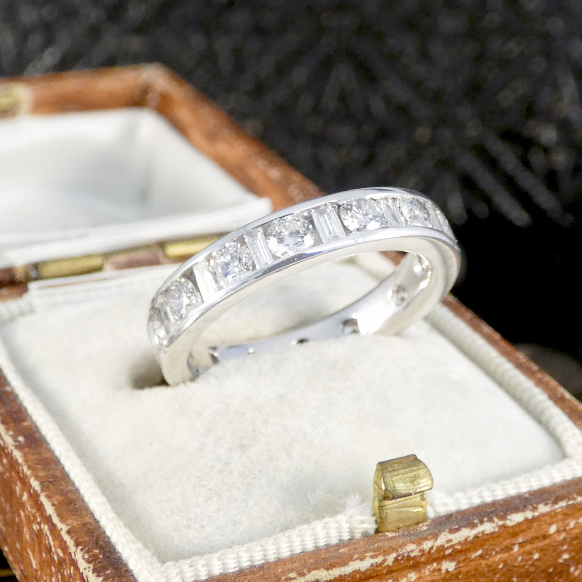 White gold eternity ring with alternating round brilliant and baguette-cut diamonds set in a channel setting, displayed in a vintage cream velvet and brown leather ring box.