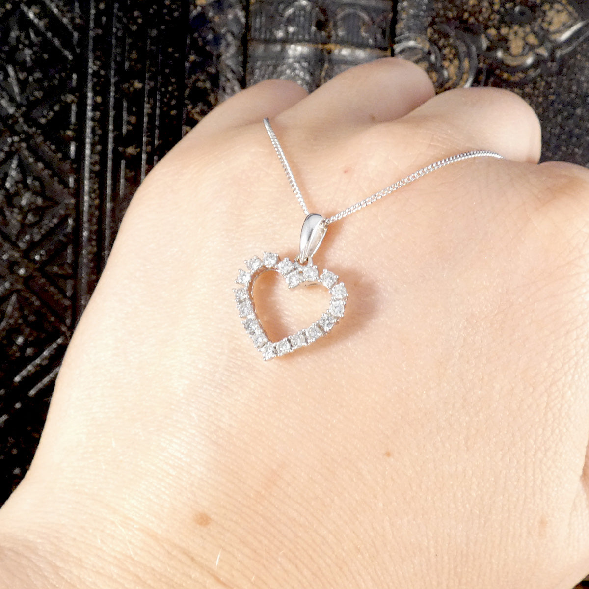 Heart-shaped diamond pendant on a chain held by a hand against a dark textured background