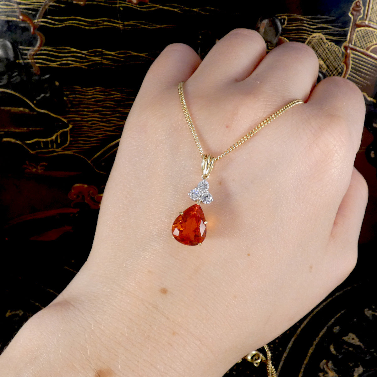 Gold necklace with a red teardrop fire opal and diamond pendant on a hand against a dark background
