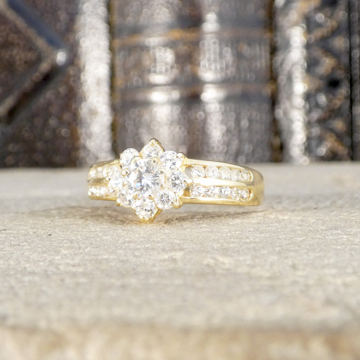 Dazzling diamond cluster ring in 18ct yellow gold with a floral-inspired design and diamond-set split shoulders, displayed on a textured stone surface.