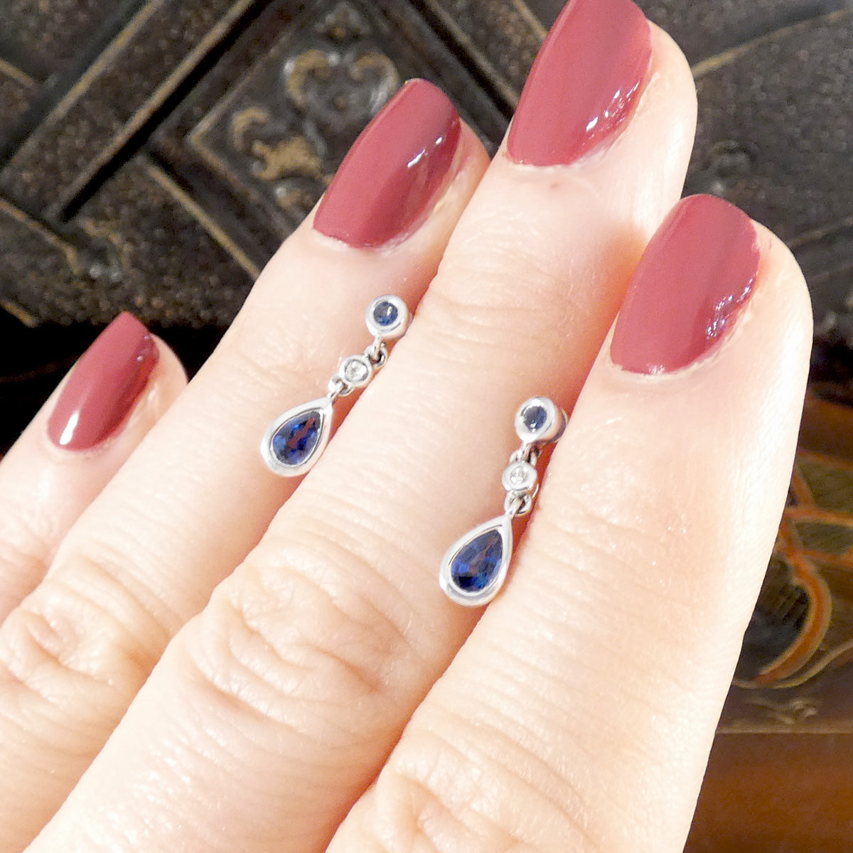 Teardrop-shaped earrings with gemstones on a hand with maroon nail polish.