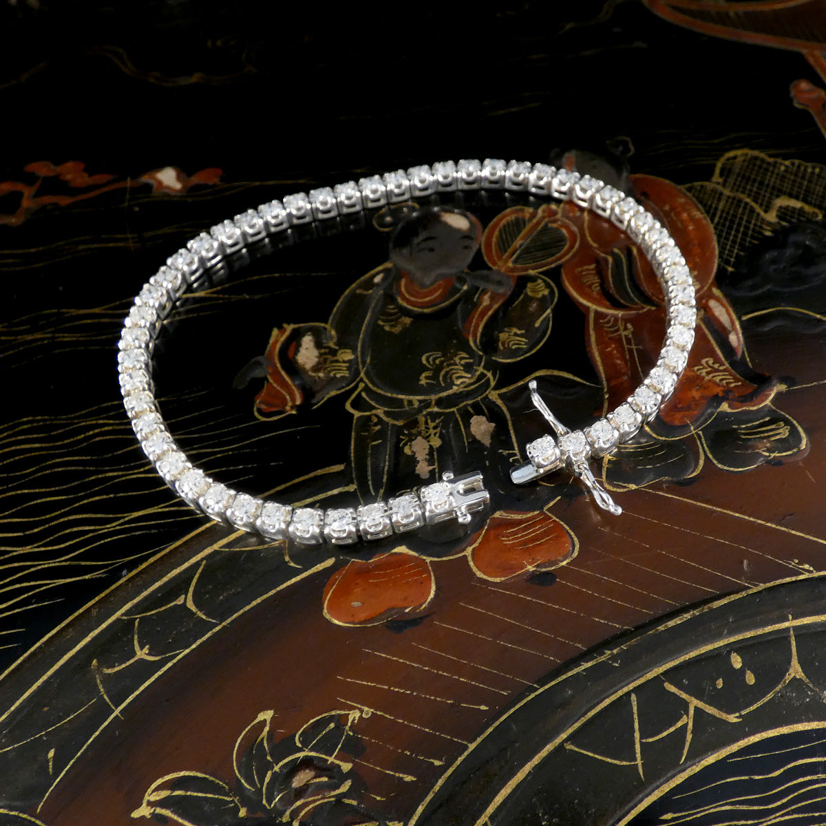 Close-up of a diamond tennis bracelet in a circular arrangement with the clasp open, showcasing round brilliant-cut diamonds set in white gold against an intricate lacquered background with painted figures. Double figure eight safety clasp.