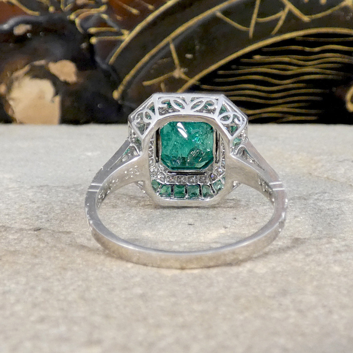Back view of Striking Art Deco-style ring featuring a central emerald-cut emerald, surrounded by a double halo of calibre-cut emeralds and pavé-set diamonds, all set in platinum. Showing open and detailed gallery setting.