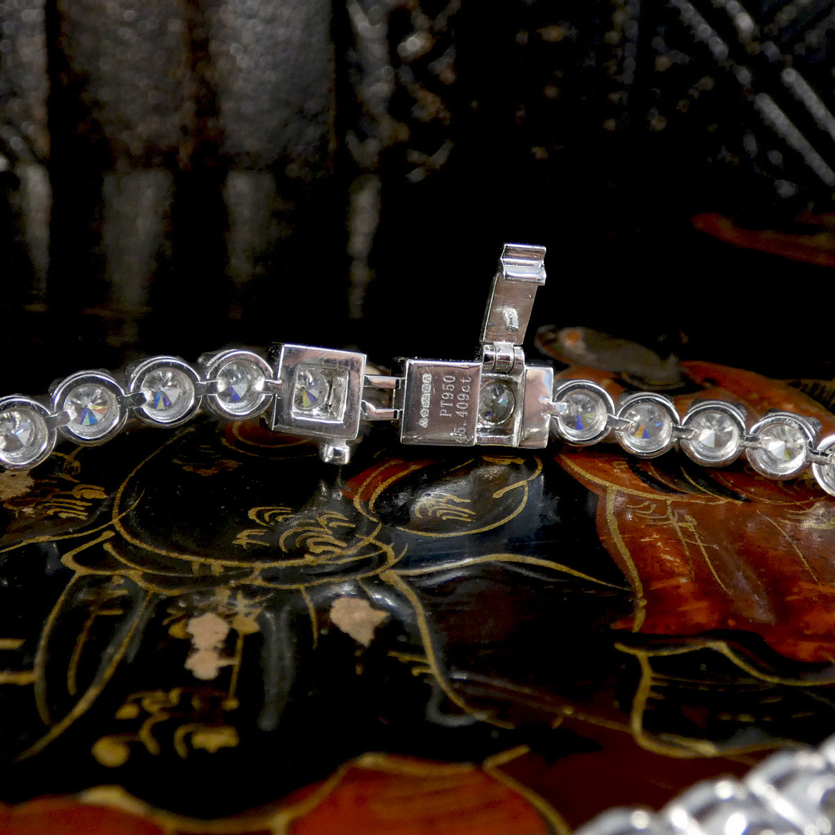 Close-up view of the clasp mechanism on a platinum diamond tennis bracelet, showcasing secure box clasp with safety latch and precision hallmark engravings, set against an ornate lacquered background.