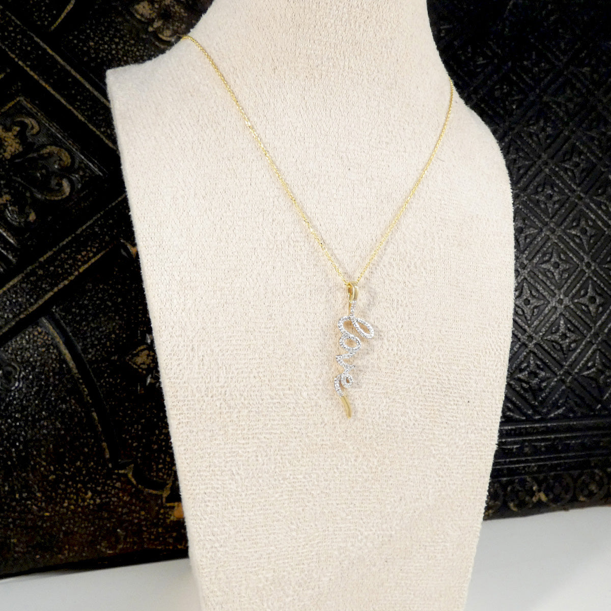 Gold necklace with a diamond set love pendant on a mannequin bust against a decorative black background.