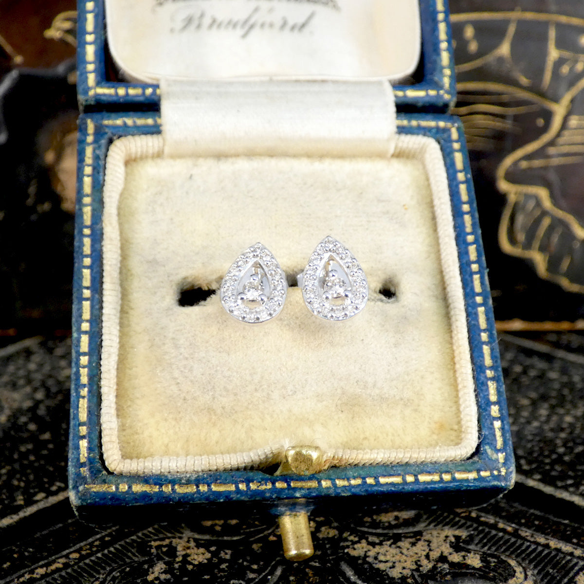 Teardrop-shaped white gold stud earrings in a decorative box on an ornate surface