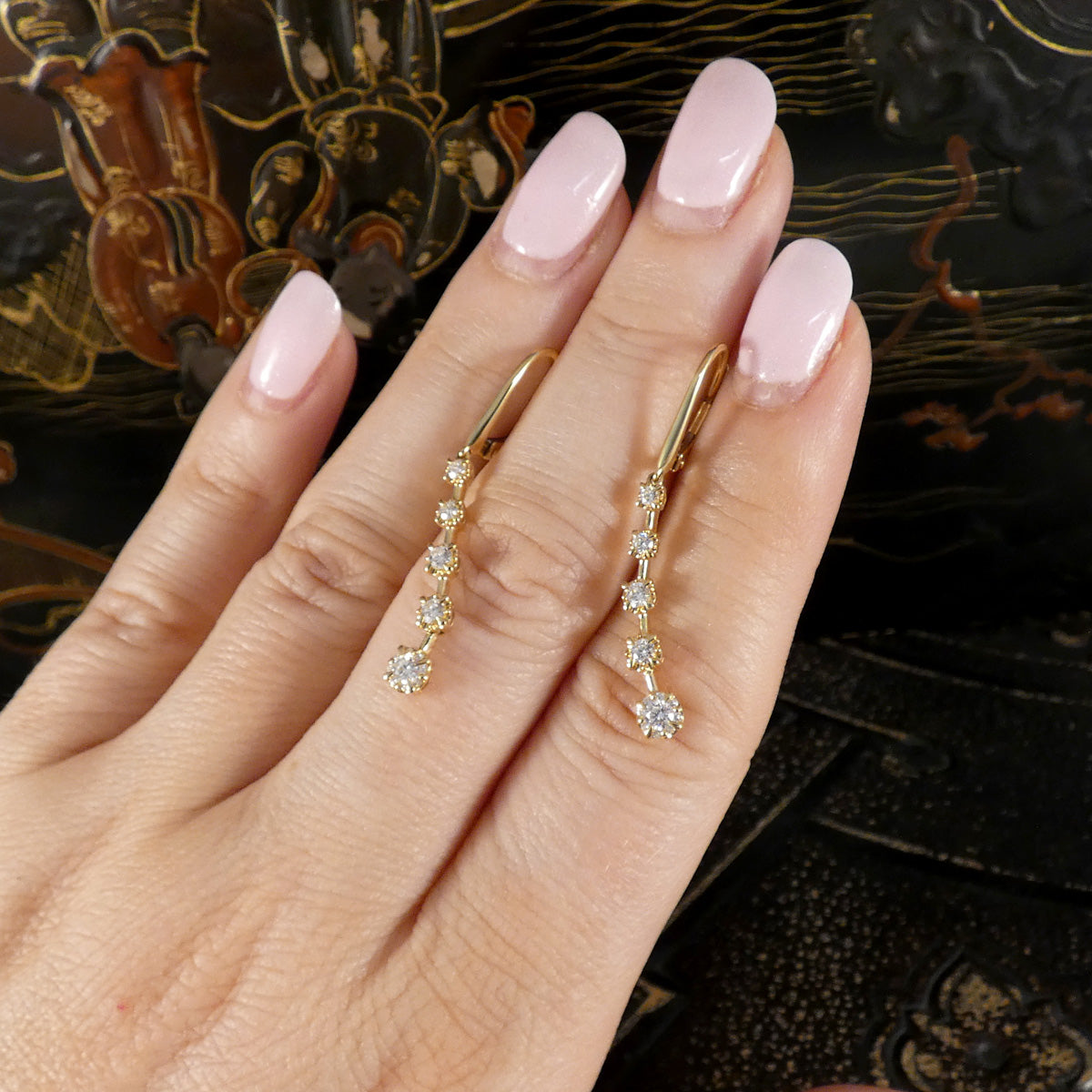 A close-up of a hand with pale pink manicured nails wearing a pair of gold drop earrings with small clear stones, set against a dark patterned background.