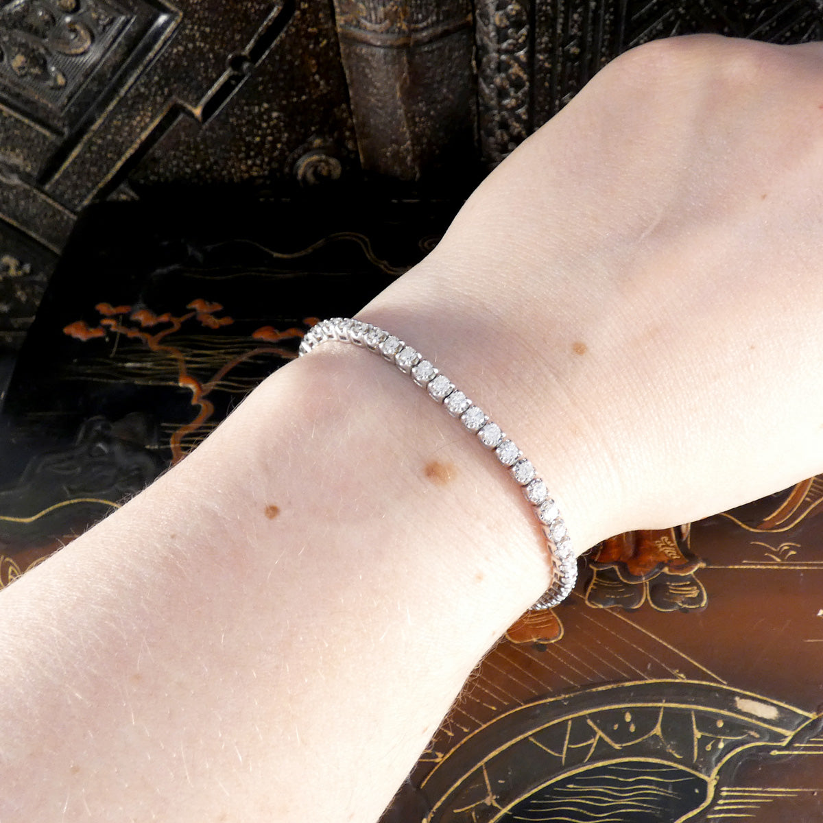 Diamond tennis bracelet worn on a wrist, showcasing a continuous row of round brilliant-cut diamonds set in white gold, with a vintage lacquered background.