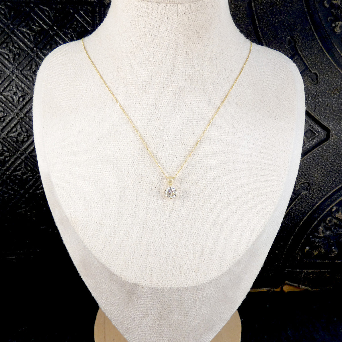 A delicate gold chain necklace with a small, clear, multi-prong gemstone pendant, displayed on a white fabric jewelry bust against a dark textured background.