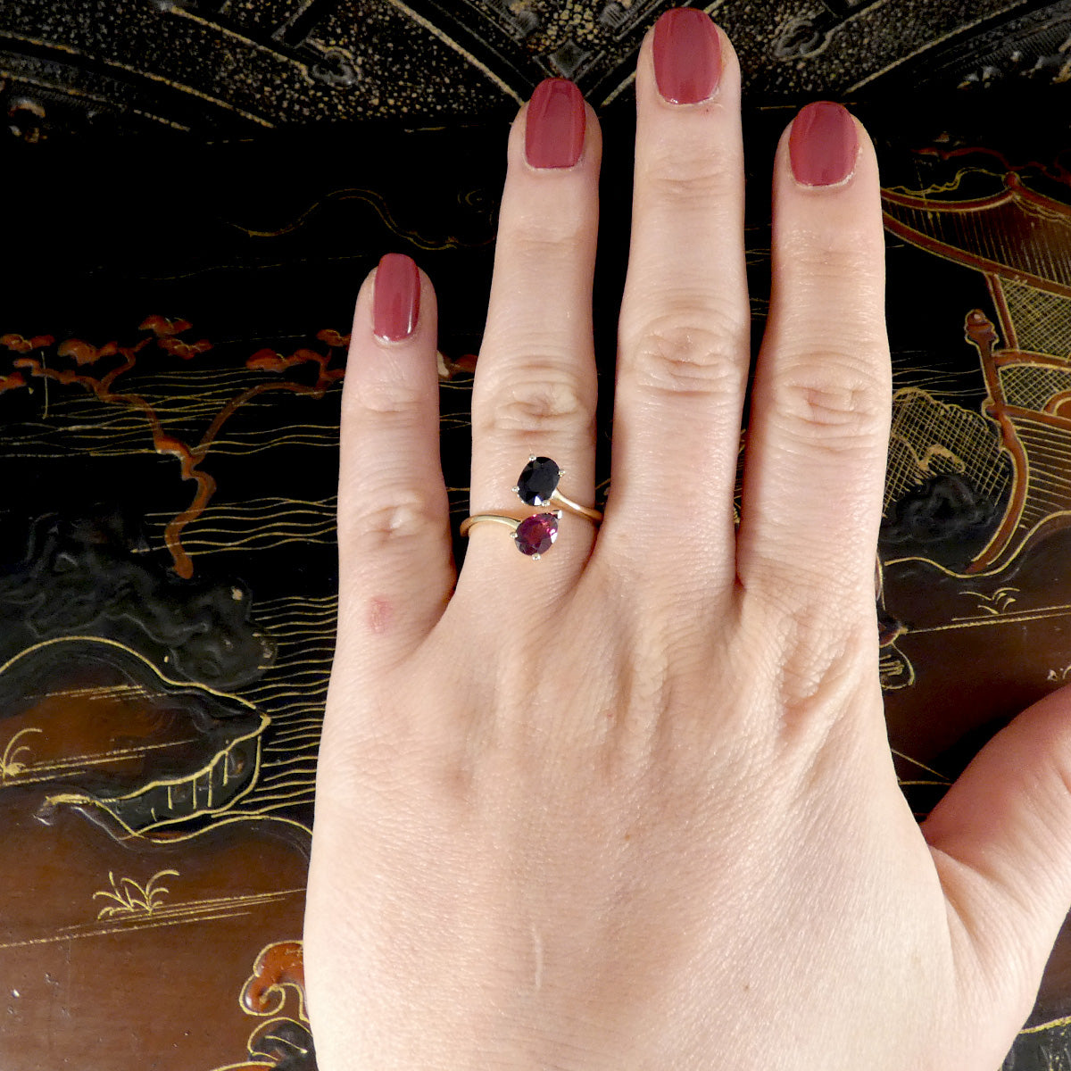 Hand wearing a gold ring with a black and red gemstone against an ornate background