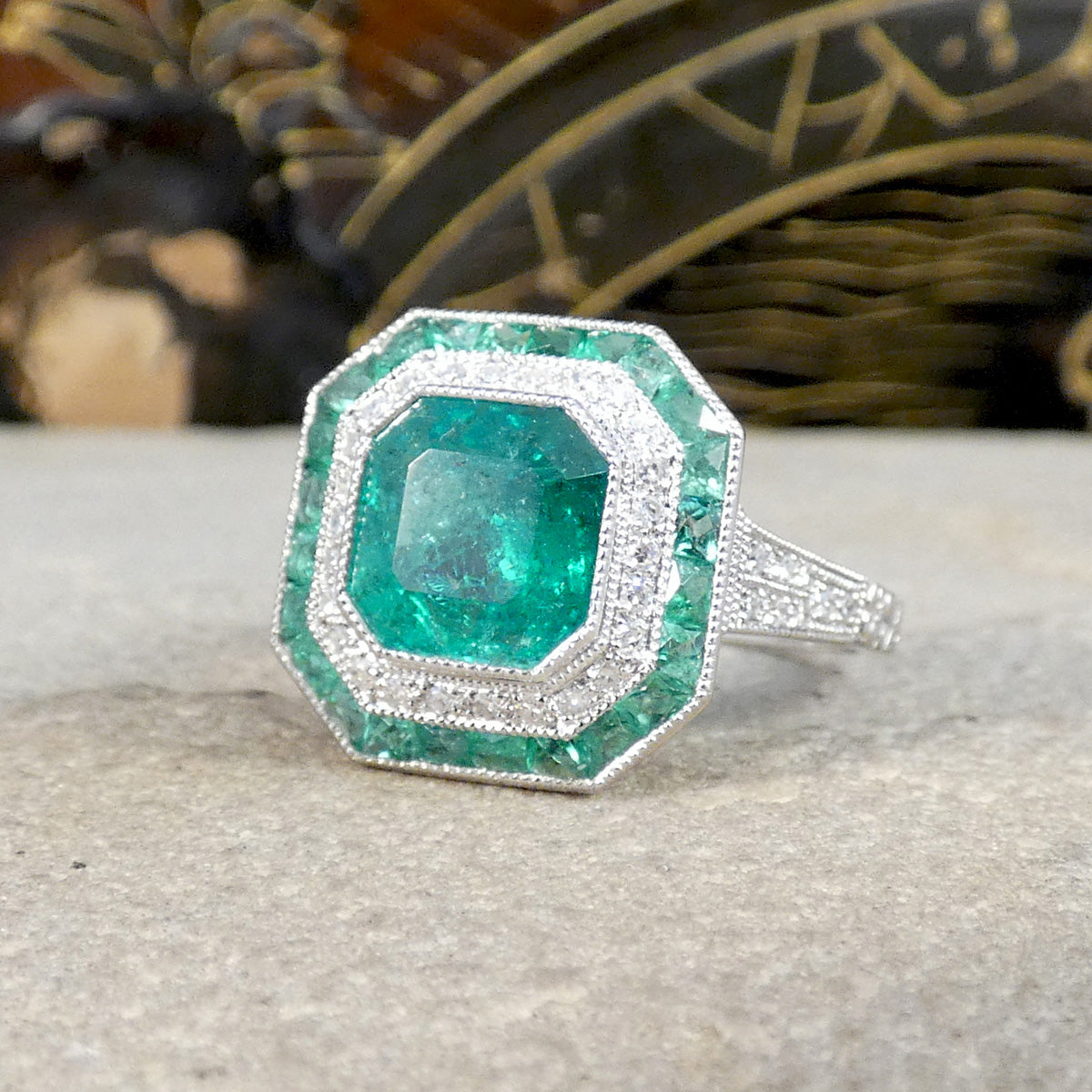 Striking Art Deco-style ring featuring a central emerald-cut emerald, surrounded by a double halo of calibre-cut emeralds and pavé-set diamonds, all set in platinum.