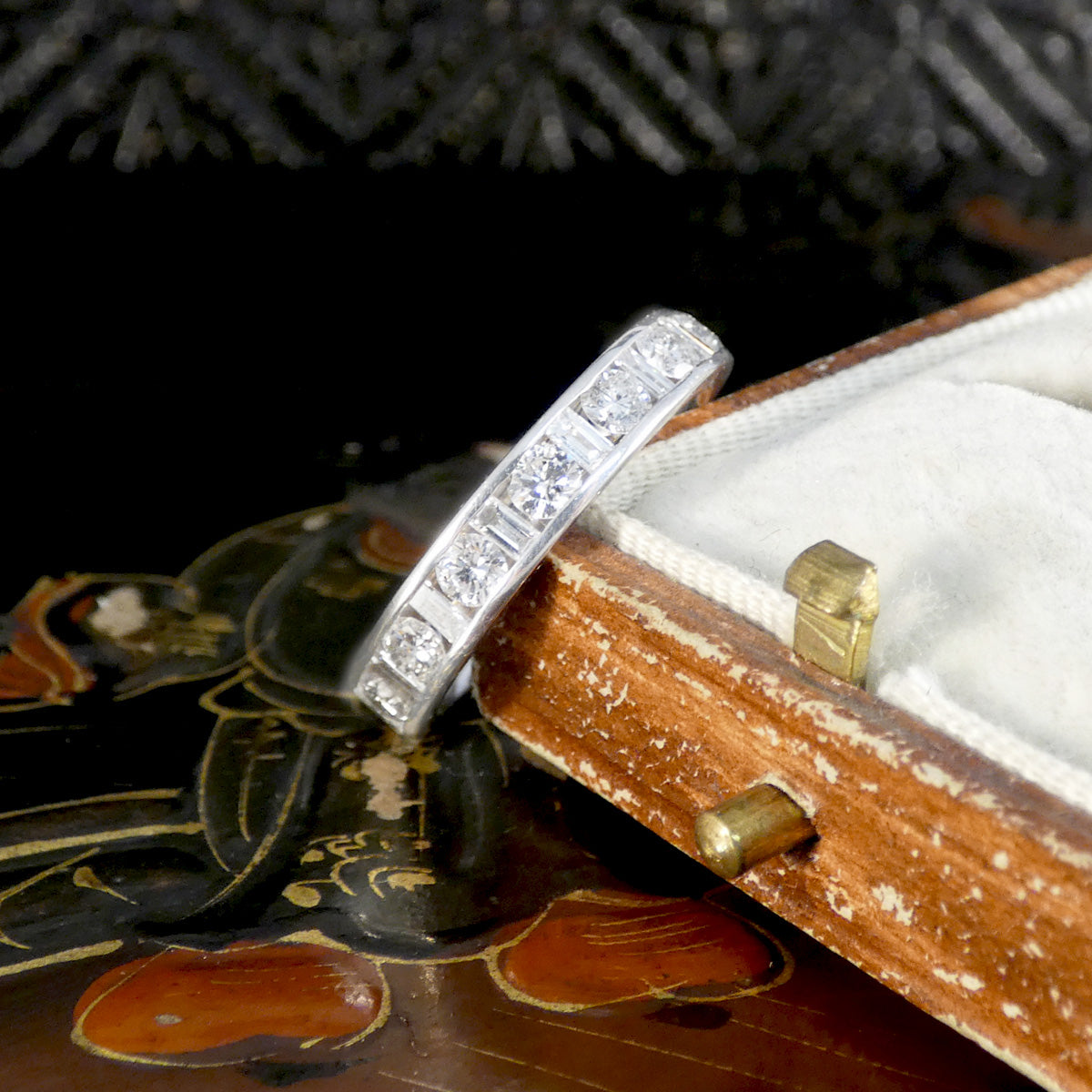 White gold eternity ring with alternating round brilliant and baguette-cut diamonds set in a channel setting, displayed against a vintage cream velvet and brown leather ring box.