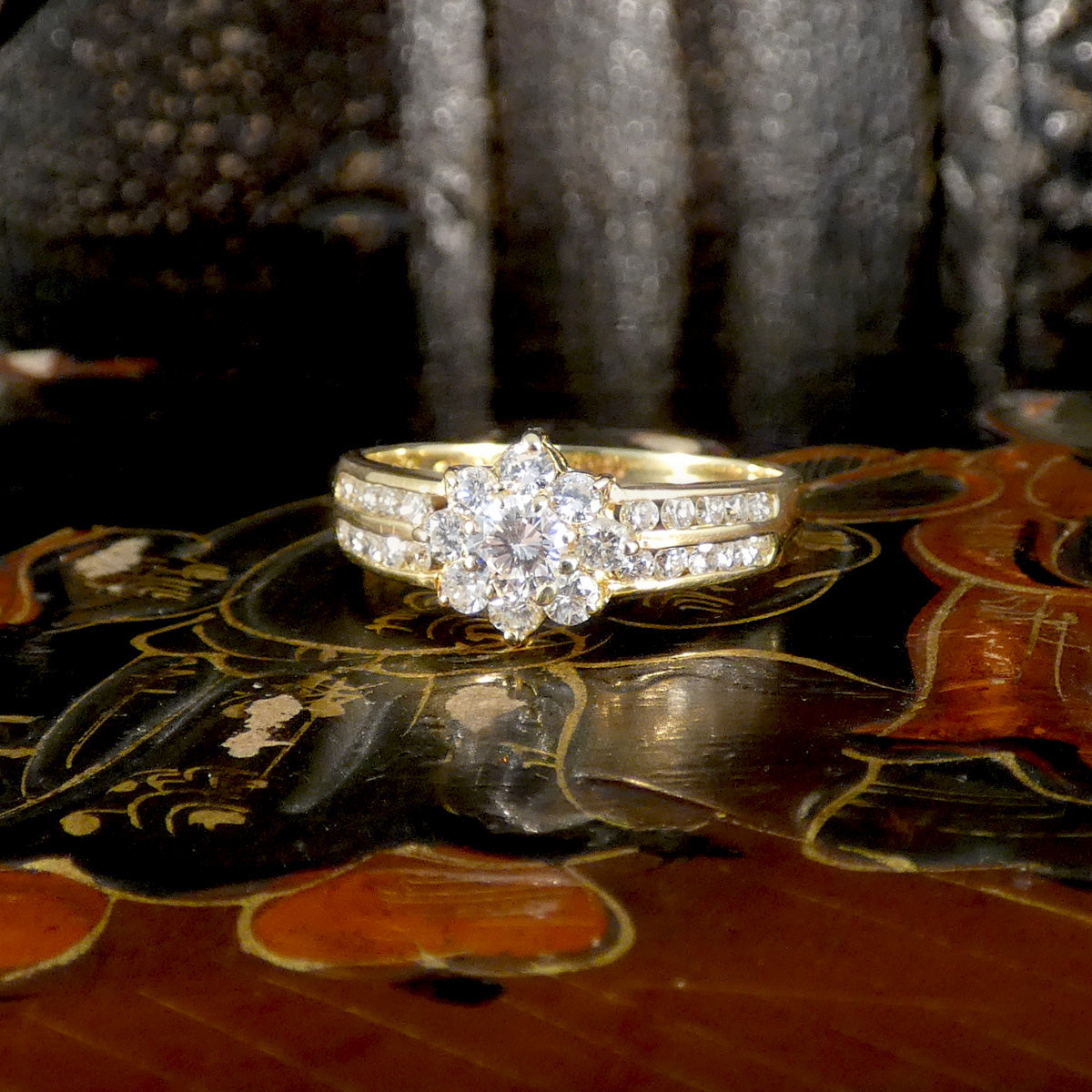 Elegant diamond cluster ring in 18ct yellow gold, featuring a round central stone surrounded by a floral halo and split shoulders adorned with accent diamonds, displayed on a lacquered oriental box background.