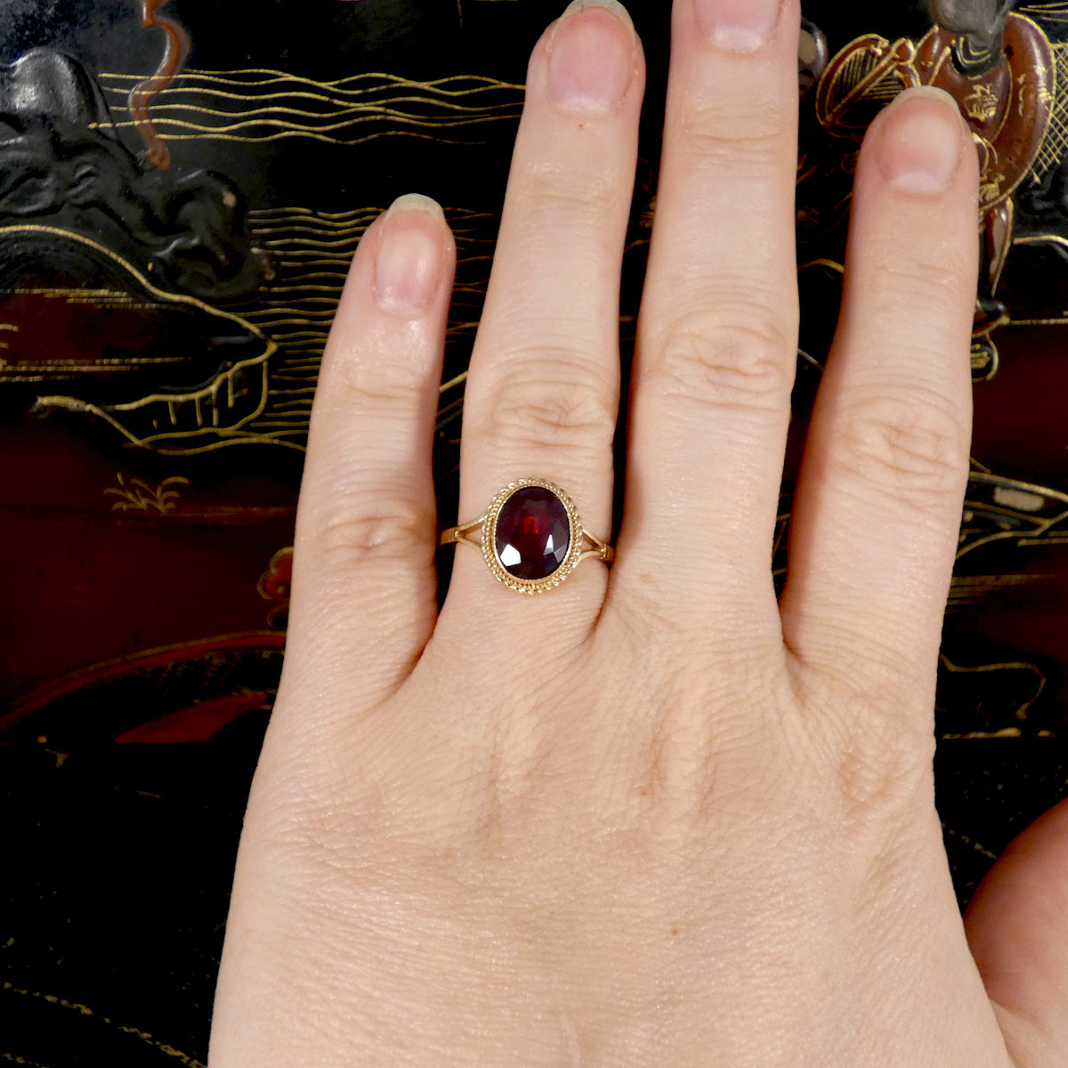 Hand wearing a gold ring with a red gemstone against a decorative background
