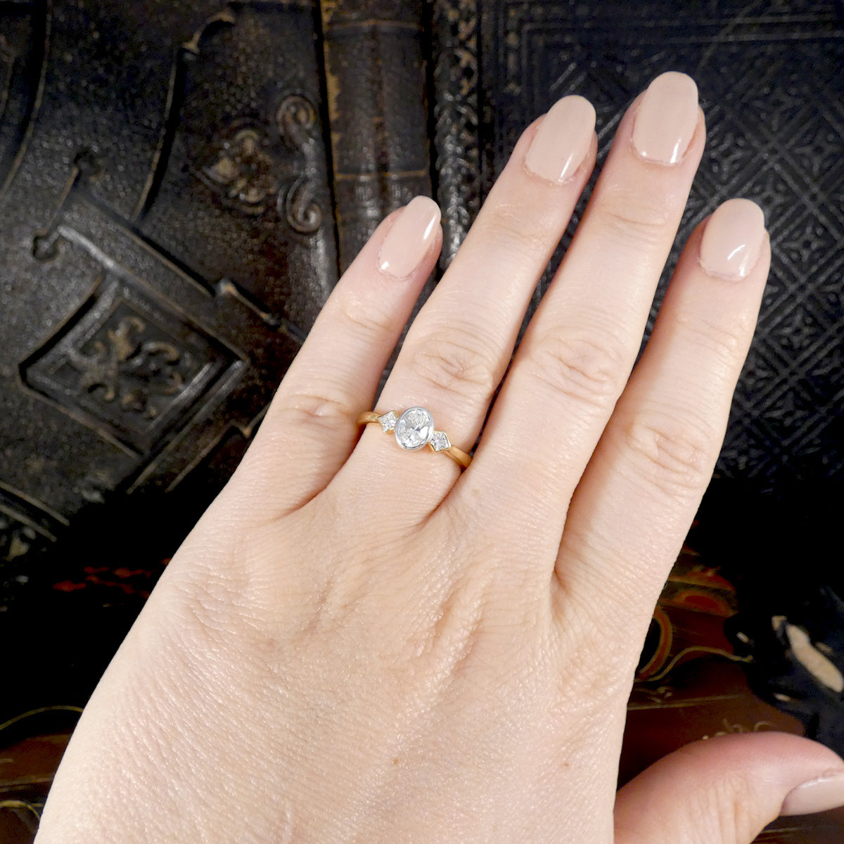 A diamond three stone ring with a central Oval cut diamond and a princess cut diamond either side fully bezel set. Displayed on the hand for side reference.