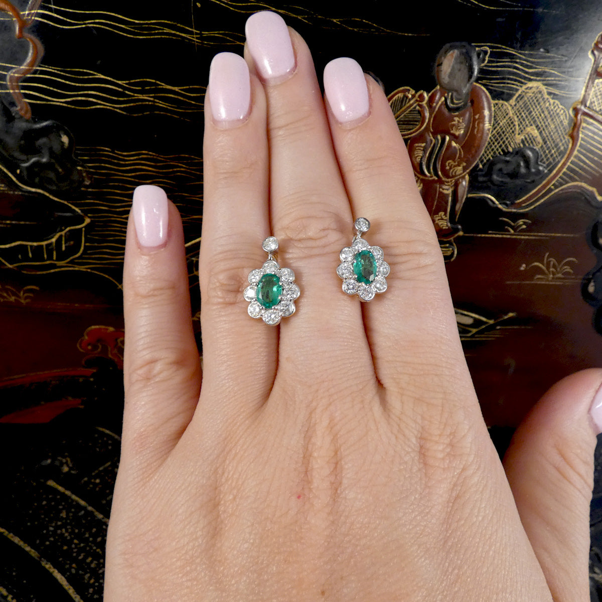 A close-up of a hand with light pink manicured nails wearing a pair of silver-toned flower-shaped earrings with oval green emerald centers and small clear stones.