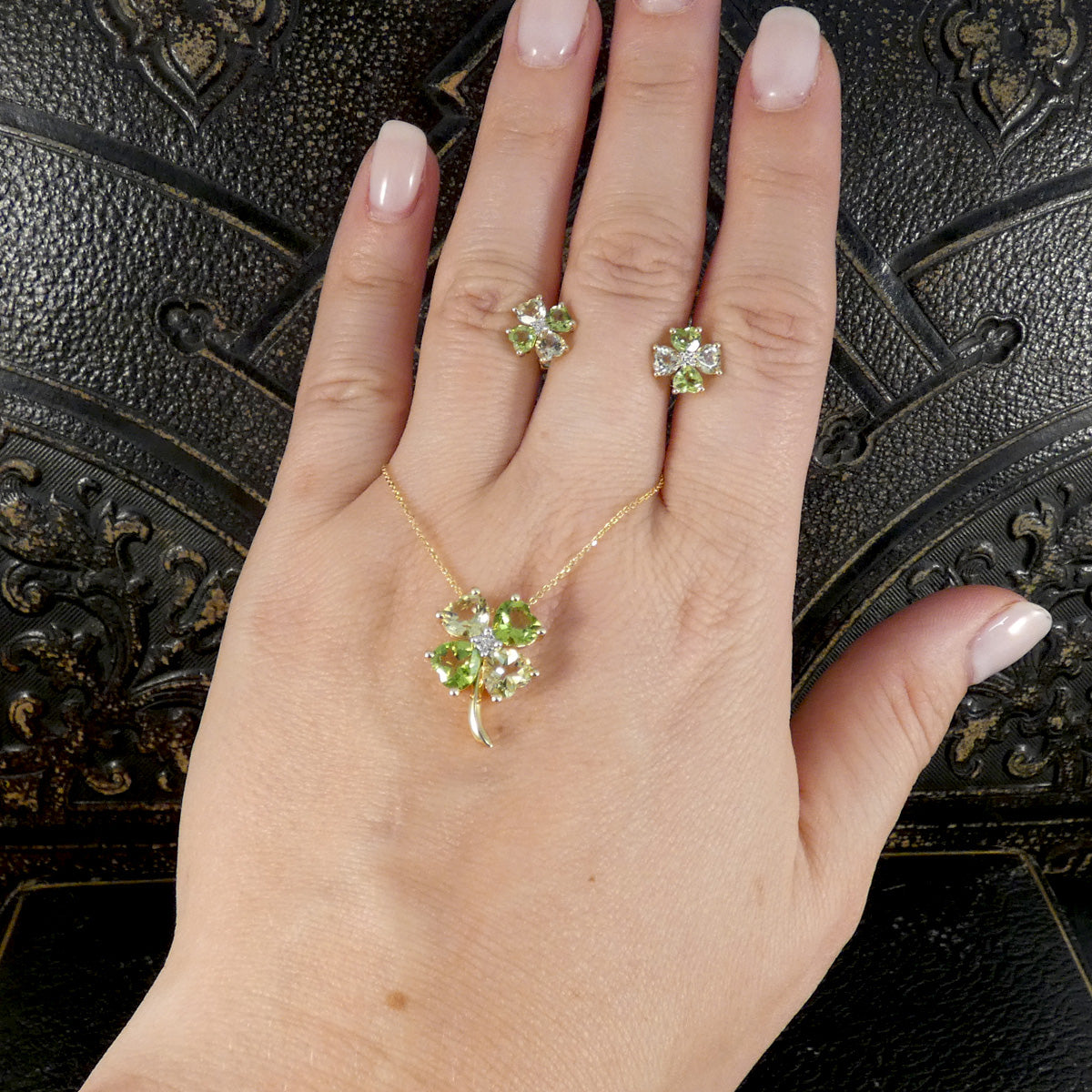 A hand wearing a gold jewelry set featuring a four-leaf clover pendant necklace, matching stud earrings, and a ring, set against a dark textured background.