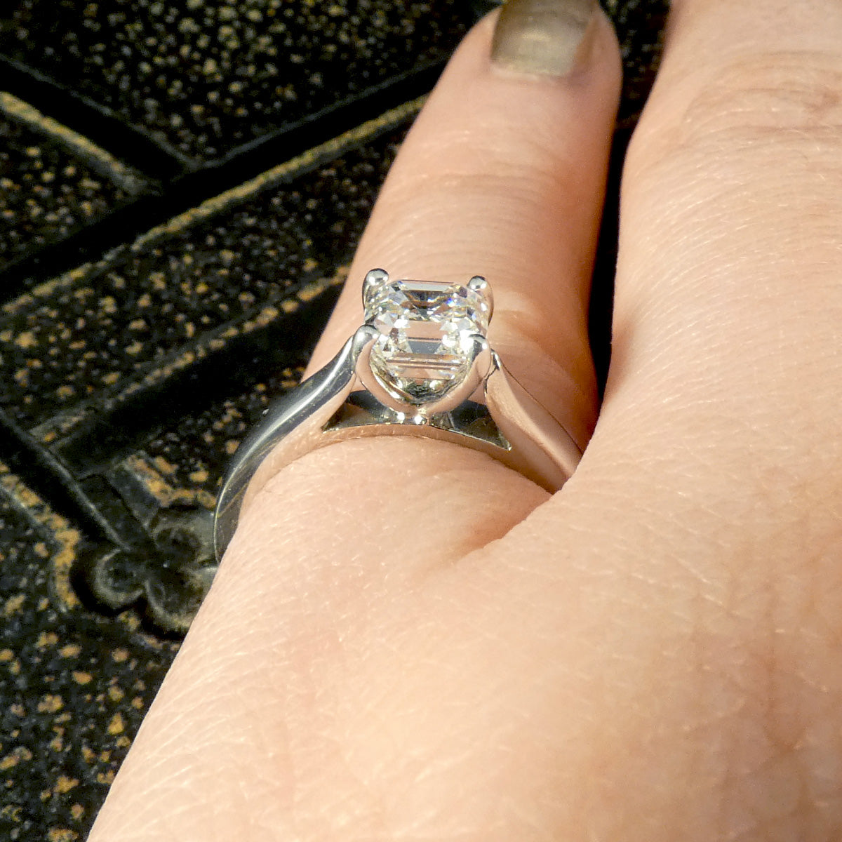 A Squared Emerald Cut Diamond Solitaire Engagement Ring in a four claw setting with a chunky Platinum band and modern feel. Shown on finger with small ring finger size and resizeable. Showing open setting allowing light o pass through.