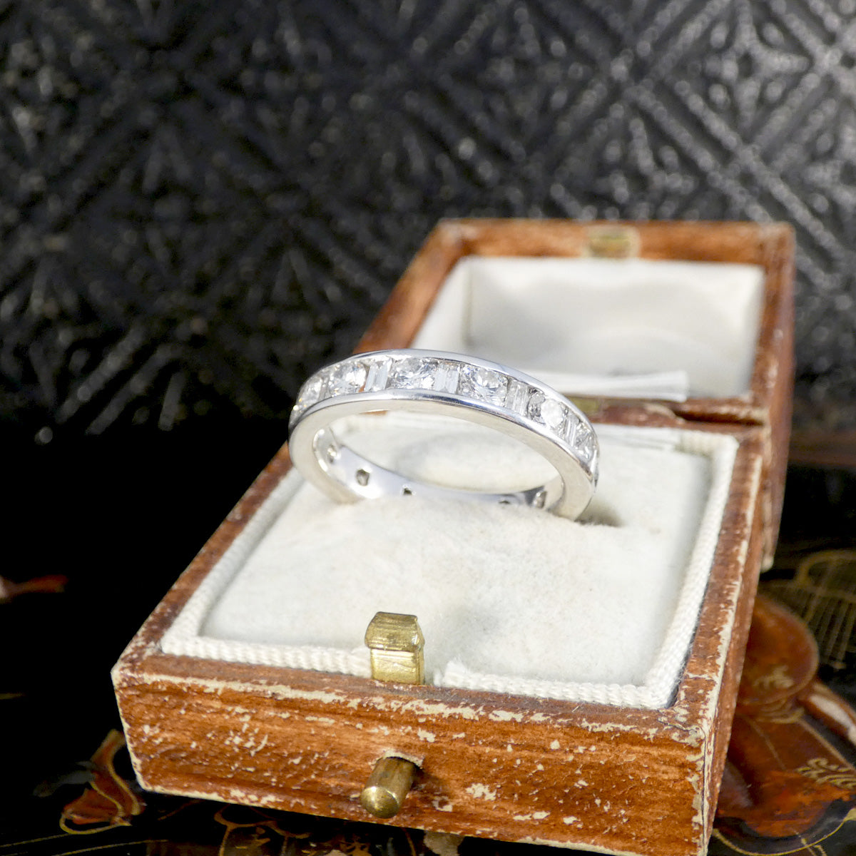 White gold eternity ring with alternating round brilliant and baguette-cut diamonds set in a channel setting, displayed in a vintage cream velvet and brown leather ring box.